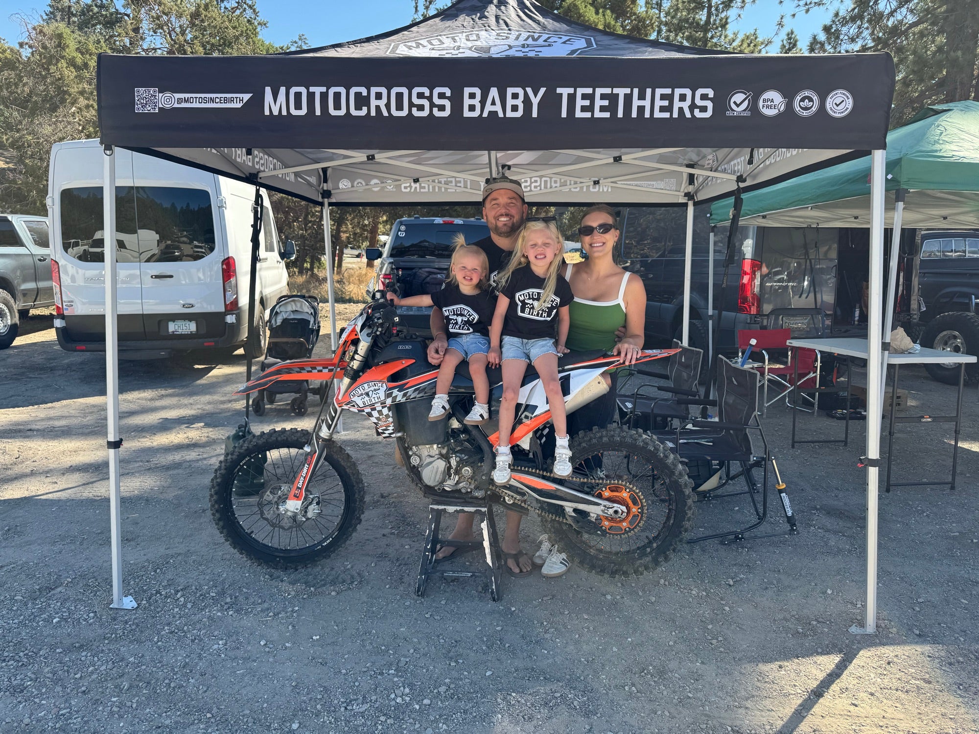 Moto Since Birth founder Mike Perlof with his family at a race, his two daughters in brand tees sitting on a dirt bike under the Moto Since Birth canopy