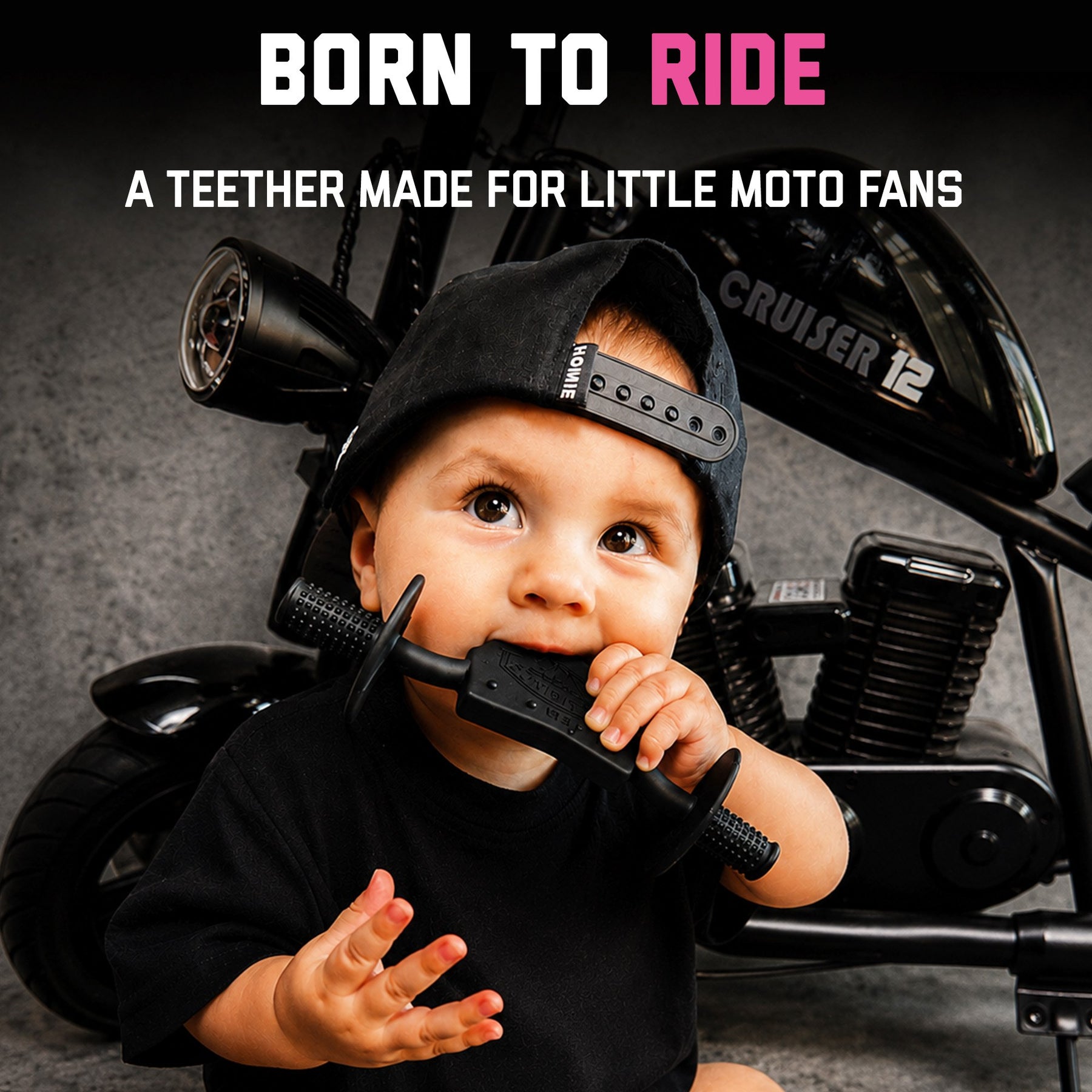 Baby in a backwards cap chewing a black Moto Since Birth teether beside a motorcycle