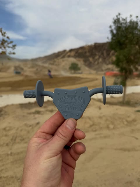 Gray Moto Since Birth teether held up over a motocross track