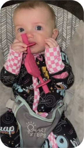 Baby in a pink checkered outfit chewing the pink Moto Since Birth teether in a bouncer seat