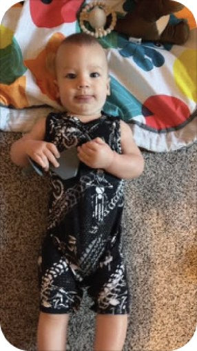 Baby in a black patterned romper on a play mat holding a Moto Since Birth teether