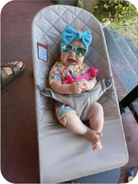 Baby in sunglasses and a teal bow holding the pink Moto Since Birth teether outdoors